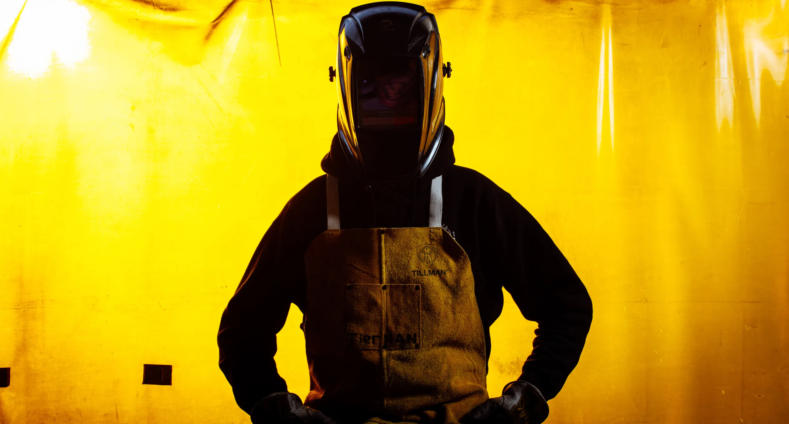 Welder wearing protective gear including a welding helmet and apron, standing against a bright yellow background. Perfect for showcasing safety standards in custom cabinet manufacturing.